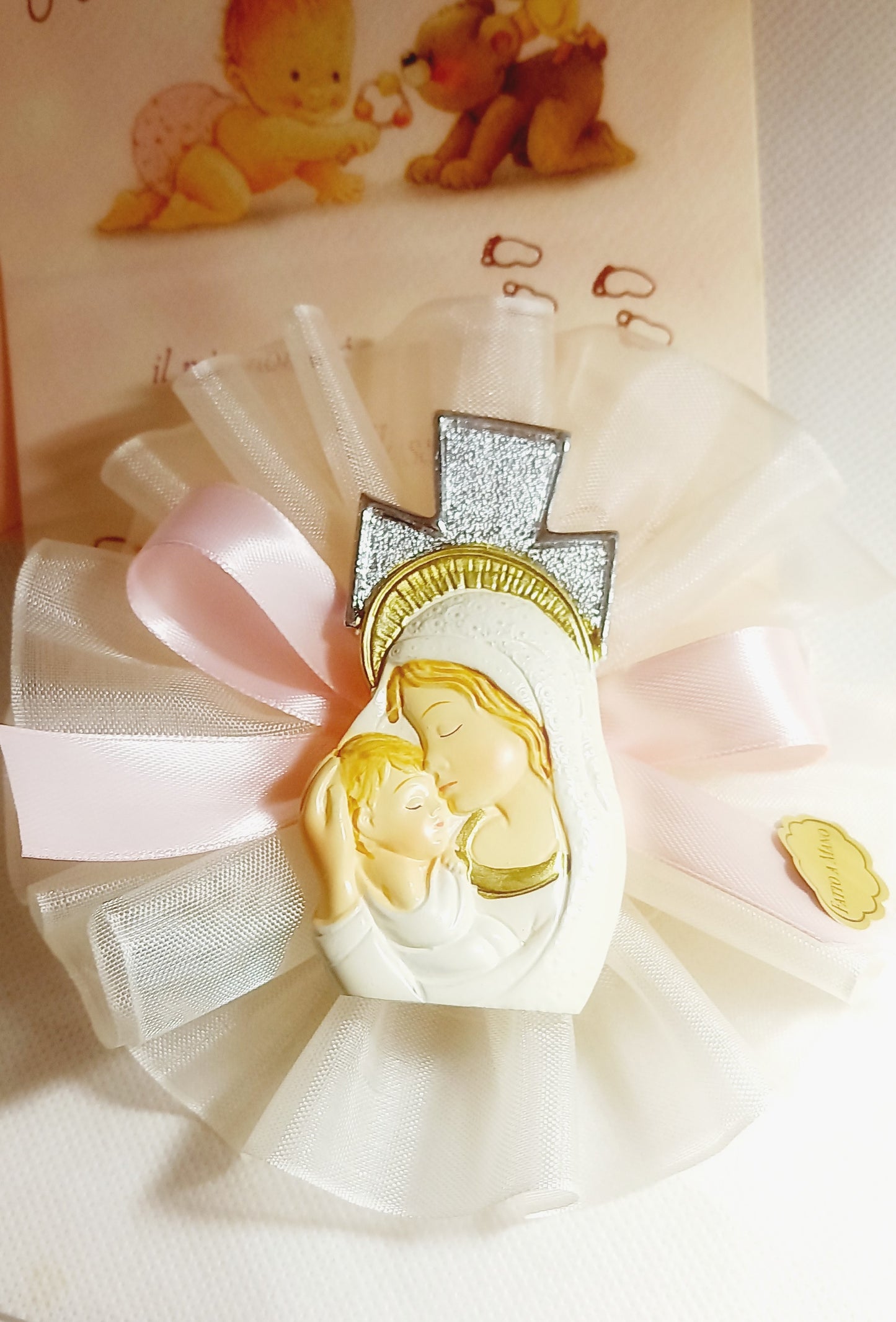 Icona in rilievo firmata Bagutta, raffigurante la Madonna con il Bambino, impreziosita da dettagli dorati e una croce glitterata argentata, decorata con una coccarda in tulle bianco e un fiocco in raso rosa. Include confetti al cioccolato e un bigliettino personalizzabile.