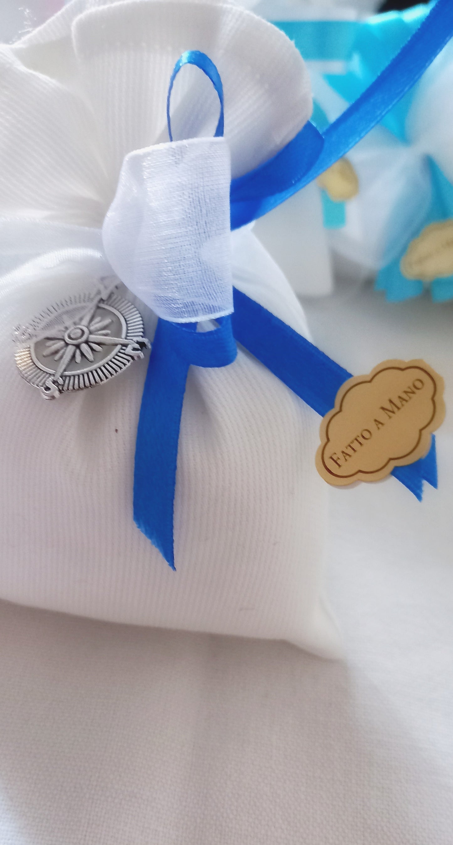 Bomboniera sofisticata con un sacchetto bianco e una Rosa dei Venti argentata, simbolo di guida. Contiene confetti al cioccolato e un bigliettino con nome e data, ideale per eventi come Comunione o Cresima. I nastri blu e bianchi possono essere personalizzati.