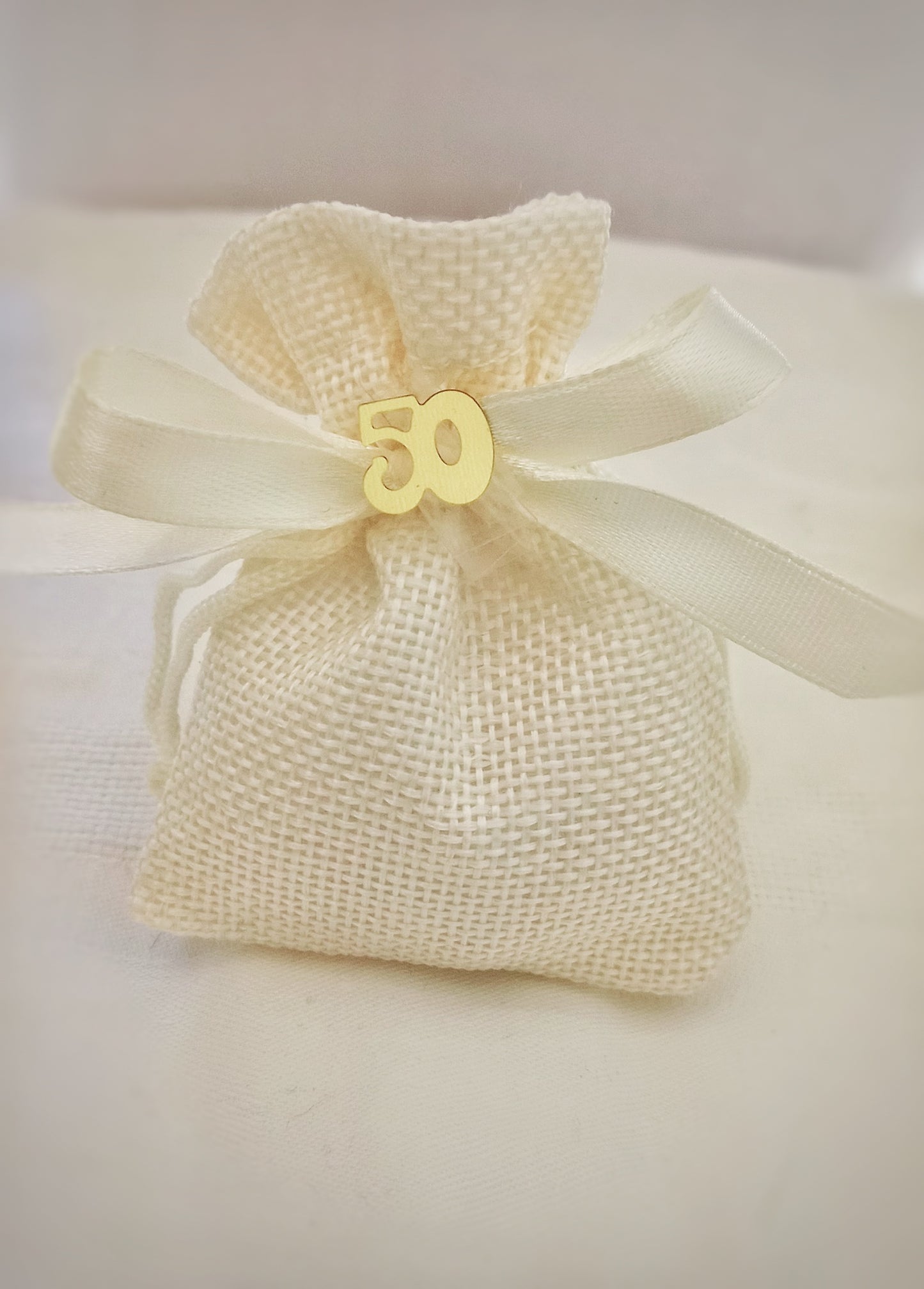 Sacchettino in juta ecru, chiuso con un nastro in raso avorio, liscio e setoso. Contiene tre confetti al cioccolato. Disponibile in altri colori e con il numero "50" dorato.