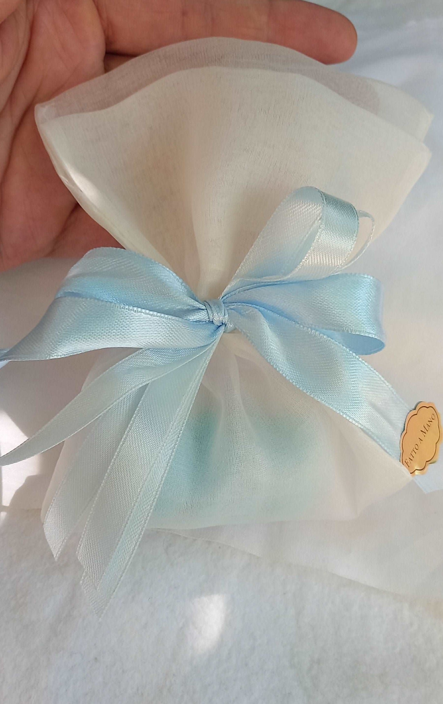 Sacchettino porta confetti in organza avorio chiuso con fiocco azzurro, etichetta "Fatto a mano " visibile sul lato.