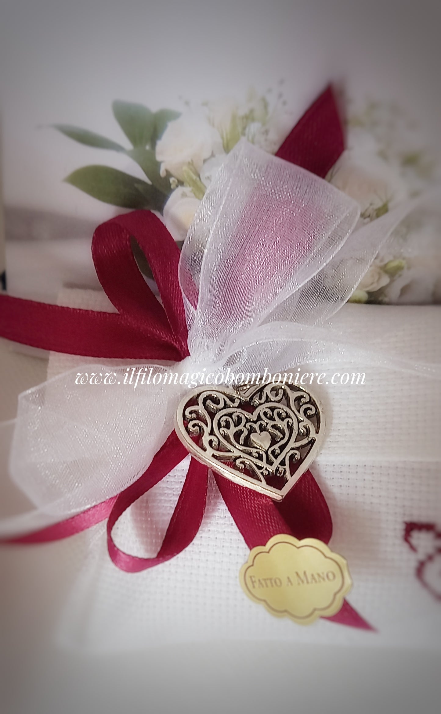 Iniziali ricamate a mano in bordeaux su una busta bianca chiusa con nastri in organza e raso abbinati a un raffinato ciondolo cuore argentato. Tradizione e stile per il tuo matrimonio.