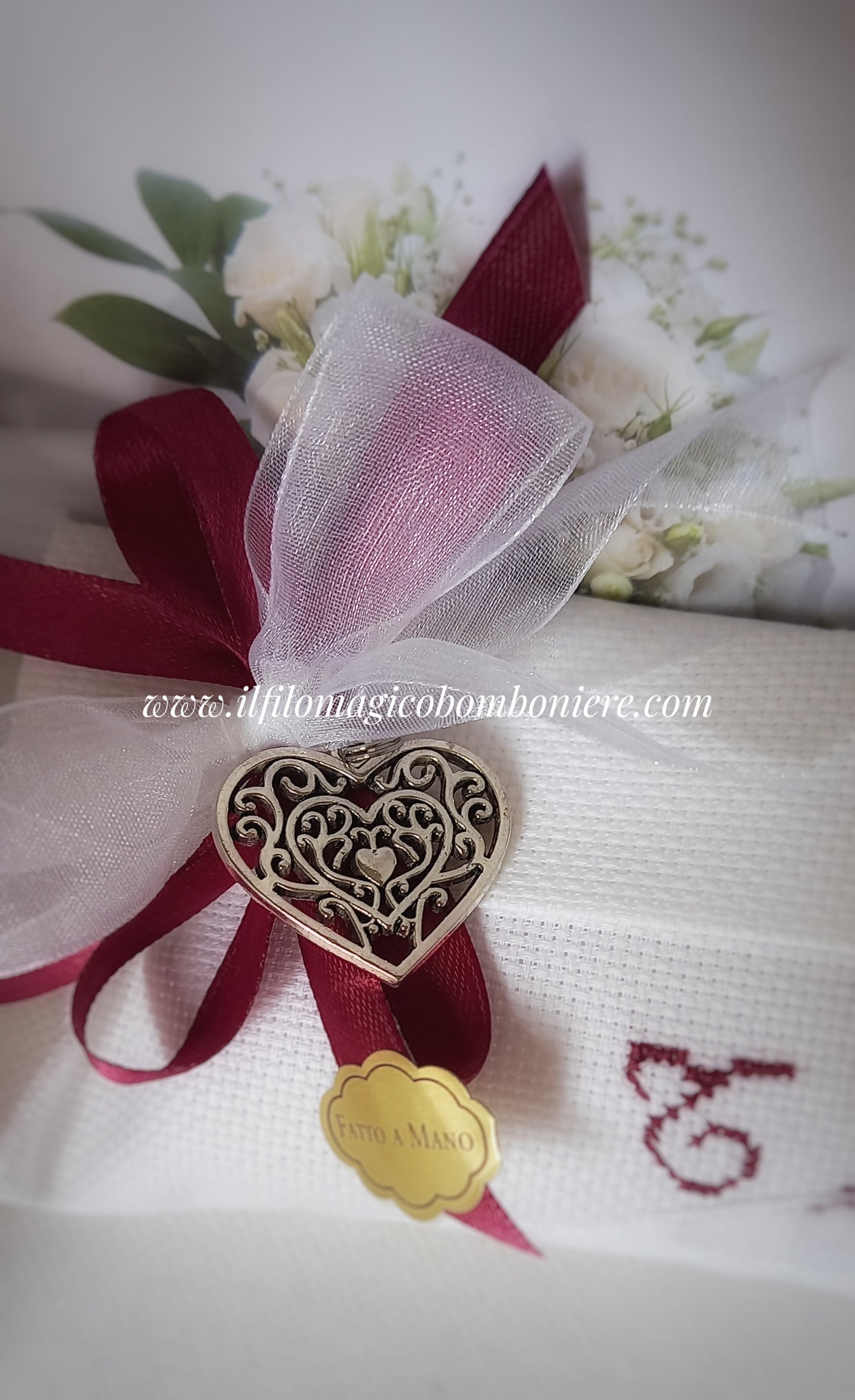 Busta bianca realizzata a mano, con le iniziali degli sposi ricamate in bordeaux. Un fiocco d'organza e raso, accompagnato da un cuore argentato, sigilla questa elegante bomboniera per un matrimonio indimenticabile.